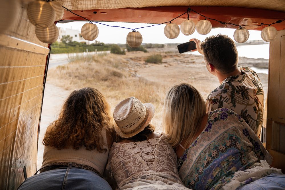 Relaxed women friends lying hanging head from trunk of car with man looking at camera