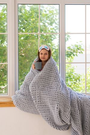 Girl wrapped in blanket by window with cup.
