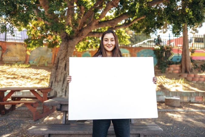 Happy teenage girl smiling at the camera while holding a blank placard in urban park