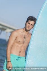 Front of shirtless male surfer standing next to blue board on the coast, vertical composition 5q88q0