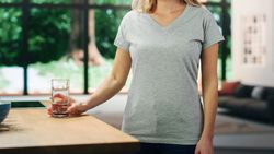 Young Adult Woman with Blond Hair Wearing Gray V-Neck T-Shirt, Holding a Glass of Water on the Table.