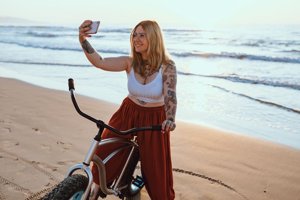 Cheerful woman taking selfie on smartphone while resting with bicycle at sunny shore