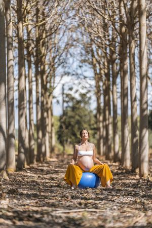 Adult pregnant lady sitting on big pilates ball in park during sunny day