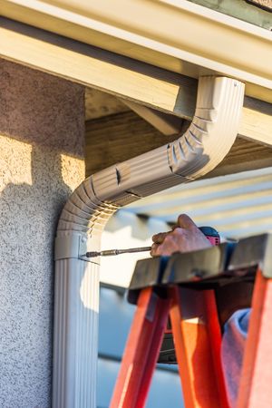 Worker Attaching Aluminum Rain Gutter and Down Spout to Fascia of House