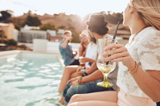 Woman holding a cocktail glass while sitting on the edge of swimming pool with friends