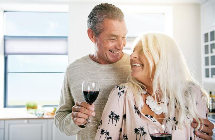 Happy older couple with red wine smiling together in a bright kitchen