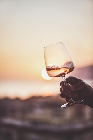 Man's hand holding glass of rose wine against sunset, vertical composition