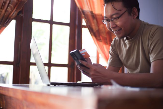 Asian Male with shirt doing online transaction in living room with credit card smartphone and laptop