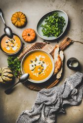 Two bowls of pumpkin soup with garnishes, bread, cream, squash, on concrete surface