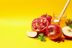 Honey pot surrounded by slices apples and pomegranate on yellow table with text for space