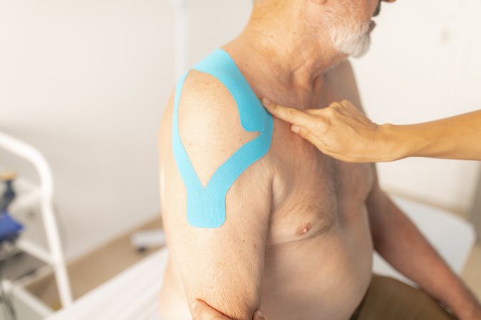 A female physiotherapist checks the tape from the shoulder of her patient, an older man