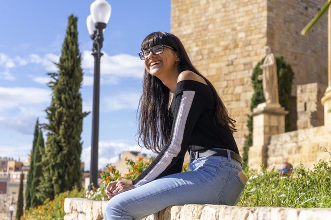 Laughing woman in eyeglasses sitting on stoned wall outside