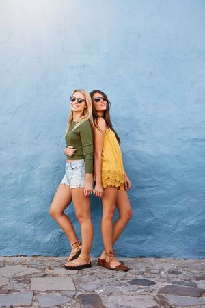 Full length shot of two women in stylish casuals posing outside against blue wall
