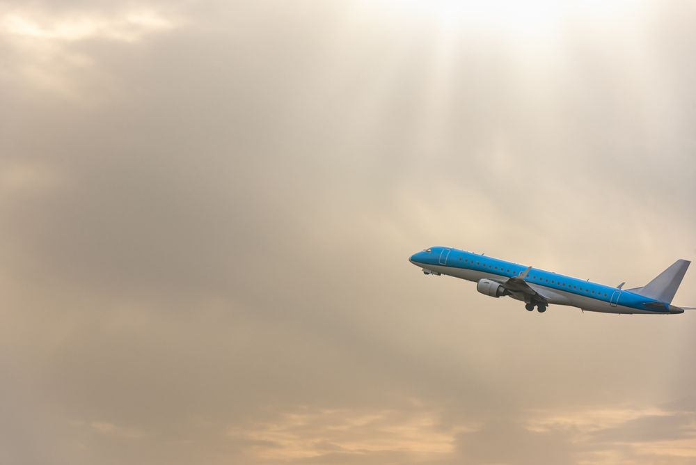 Blue airplane flying through cloudy sky with sun rays