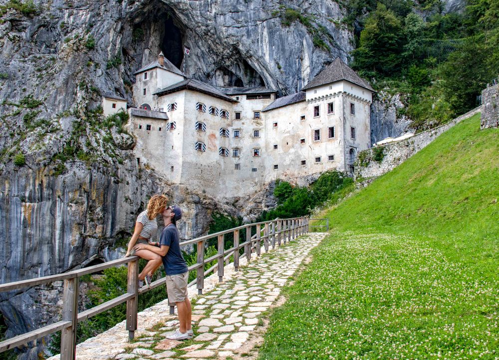 Woman and man kissing on brown wooden fence near Predjama Castle