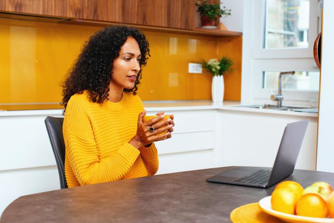 Woman in colorful kitchen sipping from mug, concentrating in front of her laptop