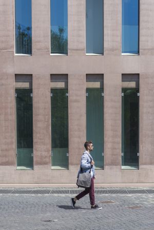 Side view of young businessman walking next to office buildings while holding a shoulder bag outdoors, vertical