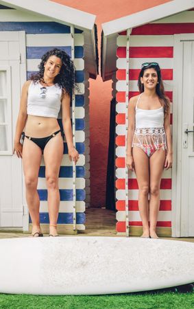 Two women in bikinis and surfboard standing in front of cabanas