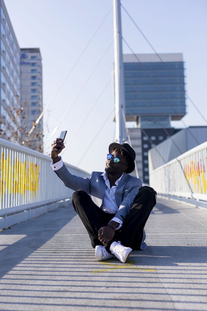 Black man in suit sitting on footbridge taking a selfie