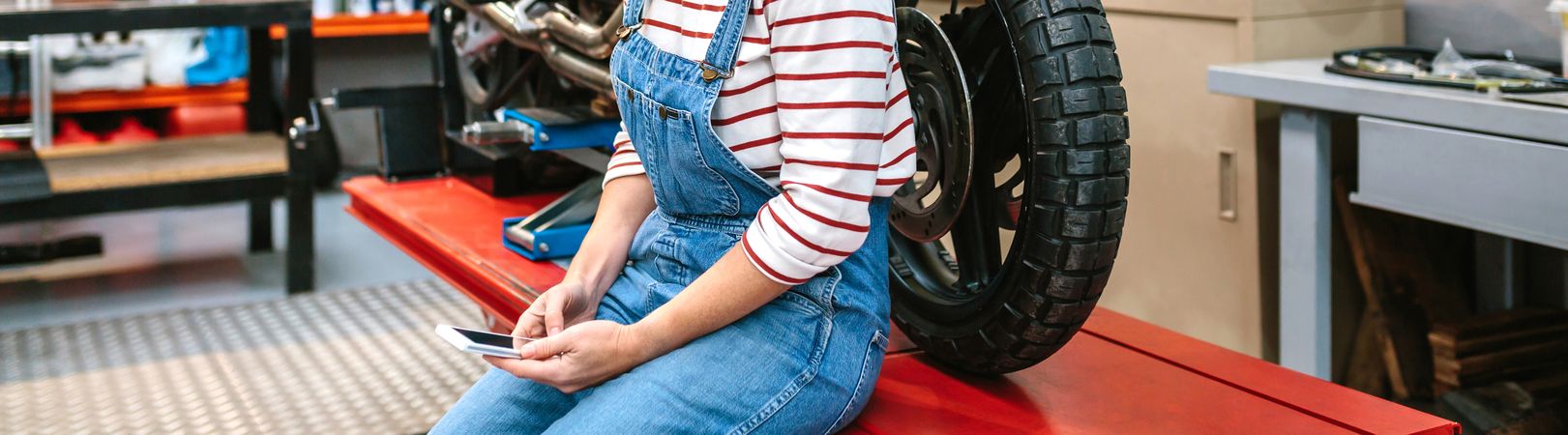 Mechanic woman looking phone sitting over platform with motorcycle on factory