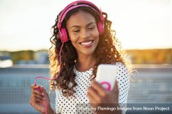 Woman in pink headphones smiling at her phone on rooftop at sunset bxLqBb