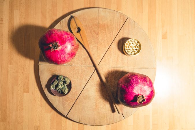 A bread board with a grinder, dried marijuana, spatula and pomegranate