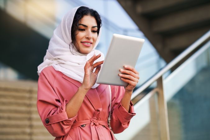 Happy female in headscarf and pink trench coat using her digital tablet