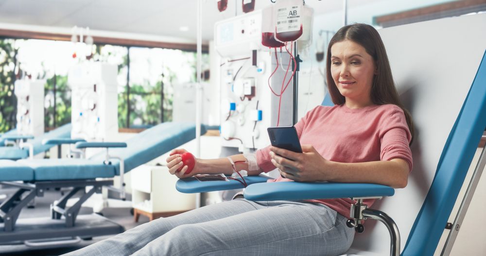 Female Donor Squeezing Heart-Shaped Red Ball To Pump Blood And Chatting Online Using Smartphone.