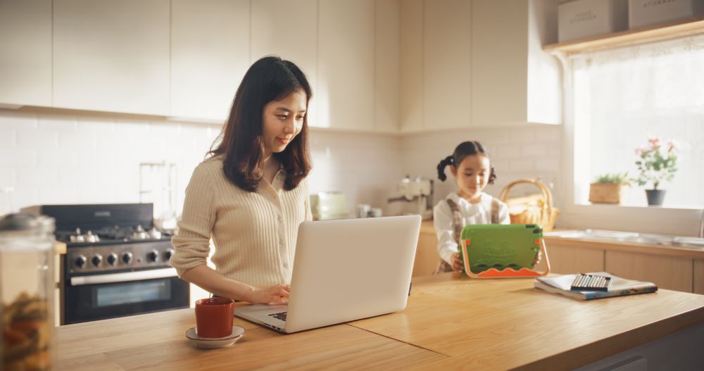 Portrait of Korean Mother Using Laptop and Smartphone To Work from Home While her Little Daughter is Using Digital Tablet.