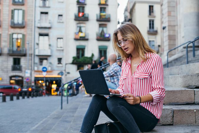 Chic woman sitting on outdoor steps in European city with open laptop
