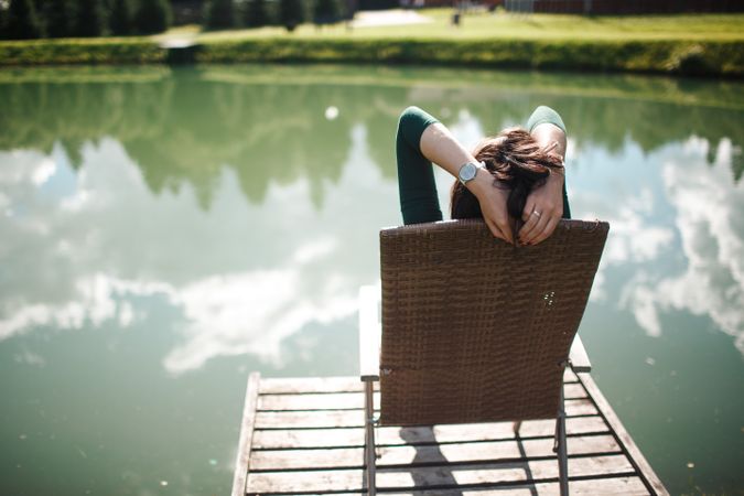 Back view of woman sitting on wooden lounge chair on a dock by lake