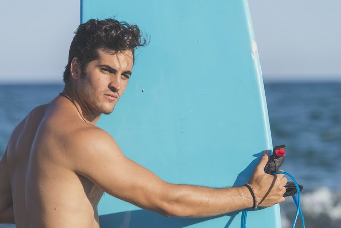 Male surfer holding blue board standing in front of the ocean and looking back at camera