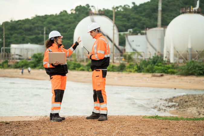 Two engineers in safety gear talking work near spherical gas storage tanks. woman is pointing forward while the man checks a laptop, indicating active discussion and site coordination in an industrial