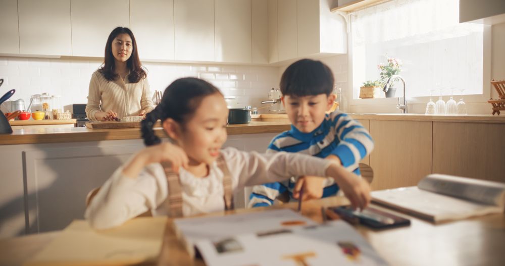 Portrait of a Korean Children Sitting at a Kitchen Table, Drawing, Writing While Mother is Preparing Breakfast.