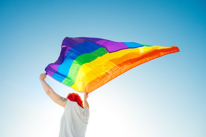 Young woman in light top waving rainbow flag under blue sky