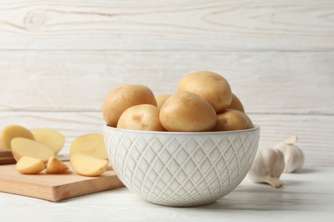 Side view of bowl full of potatoes on kitchen counter with garlic and cutting board, copy space