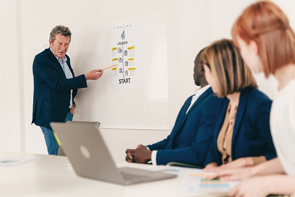 A mature man presents a project timeline on a wall panel in a modern office in front of colleagues