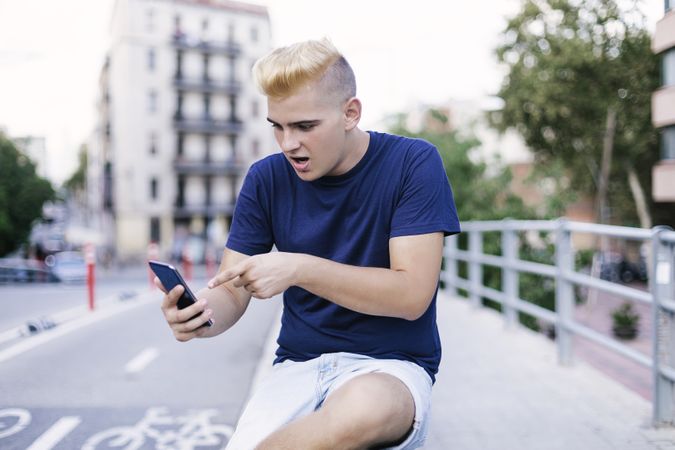 Young man with blonde hair sitting on the street surprised by something on a mobile phone
