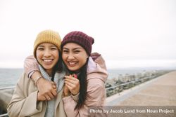 Two happy young Asian women in beanies embracing 47el60