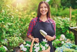 Confident woman pictured in a greenhouse holding a basket of flowers and holding a a trowel