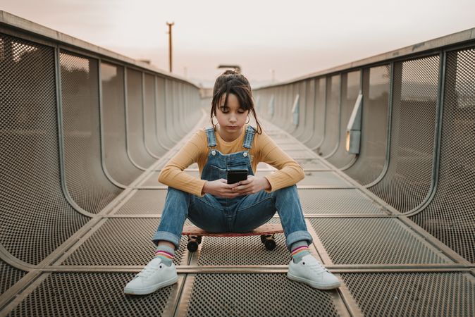 Girl with smartphone sitting on skateboard on walkway