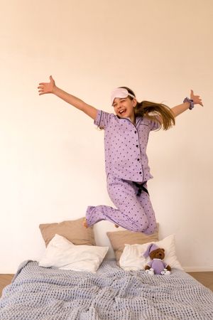 Joyful girl jumping on bed in purple pajamas.