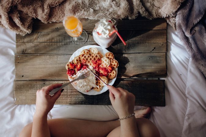 Young woman eating appetizing waffles and tasty drinks on tray on bed