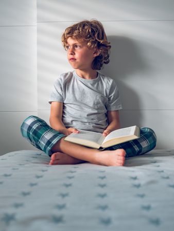 Pensive little boy sitting on bed with opened book