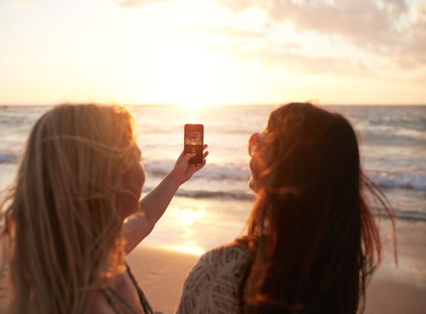 Two young women with mobile phone taking pictures of a sunset at the ocean