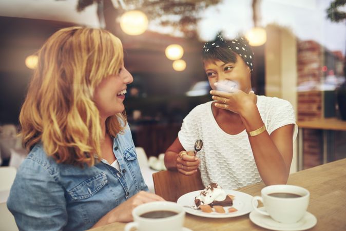 Two female friends sitting at a cafe table laughing with coffee and dessert