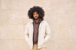 Young man with curly afro hair, dressed in jacket, hands in pockets, leaning against a wall