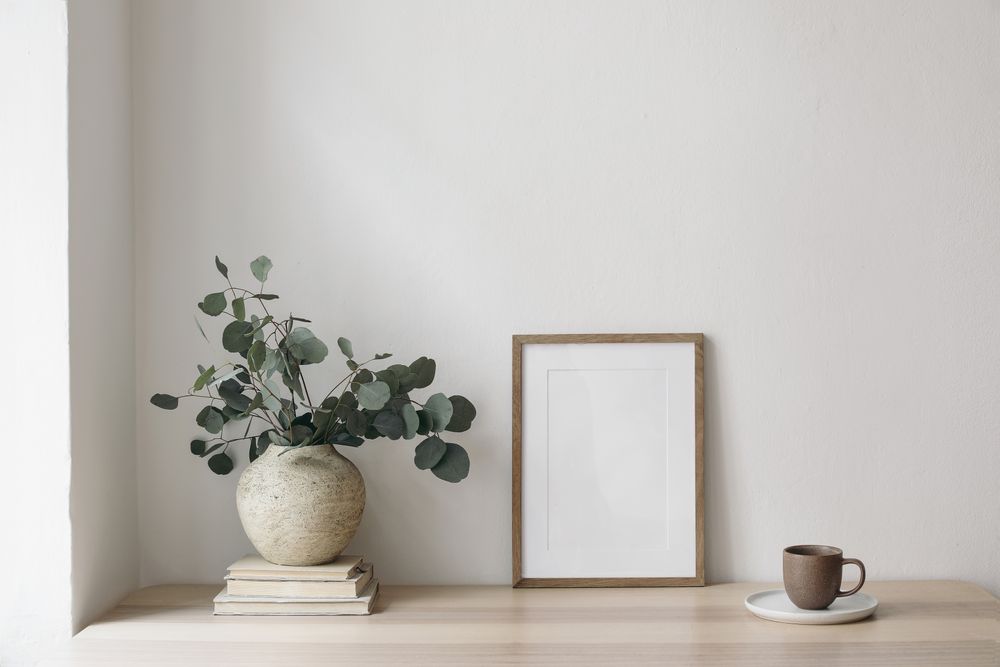 Table with vase, eucalyptus tree branches, old books on wooden table on rattan chair near window with poster mockup