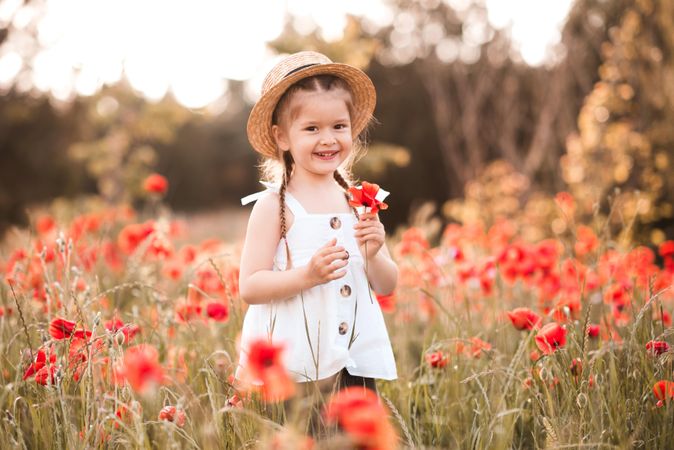 Smiling girl in light dress with hat holding red poppy flower in meadow outdoor