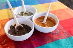 Three bowls with red, green and yellow mole sauce with a wooden spoon on top of a colorful traditional tablecloth